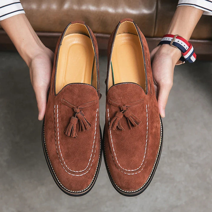 A person holds a pair of brown tassel loafers with white stitching, showing the shoes from above. Featuring durable construction and tan insoles, these loafers are positioned over a gray floor.