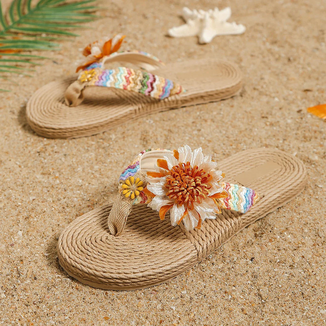 A pair of beige woven sandals with colorful straps and large fabric flowers, designed as a women's fashion accessory, rests on sandy beach with a seashell and a palm leaf nearby.