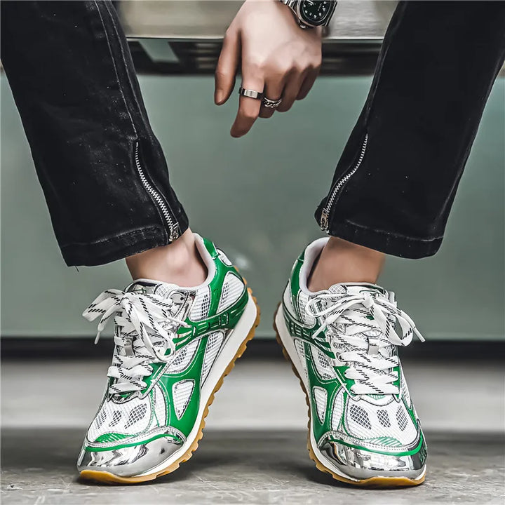 A person wearing black pants with zipper details and green, white, and silver lightweight mesh shoes is seated, showing off the shoes’ cushioned insole and non-slip outsole along with rings on their fingers and a watch on their wrist.