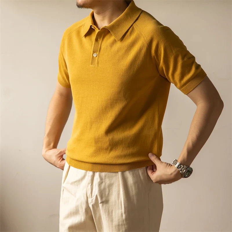A man wearing a mustard yellow mens polo shirt and beige pants stands against a plain light background, with his arms slightly bent and his hands resting at his hips, showcasing a classic style polo look.