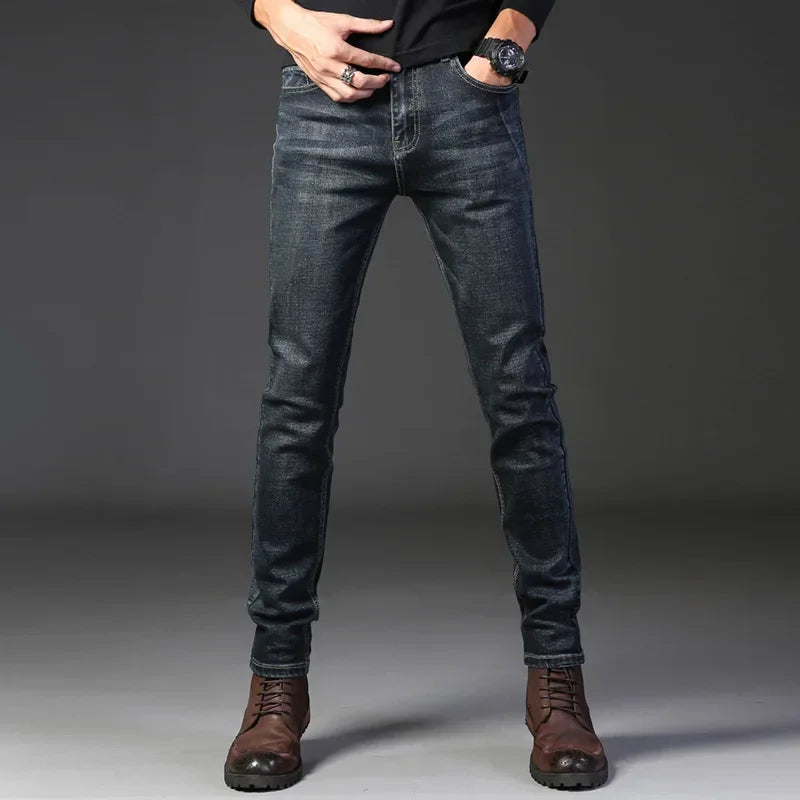 A person wearing dark slim-fit jeans crafted from stretch denim, a black long-sleeved top, brown lace-up boots, and a watch poses with hands in their pockets against a plain dark background.