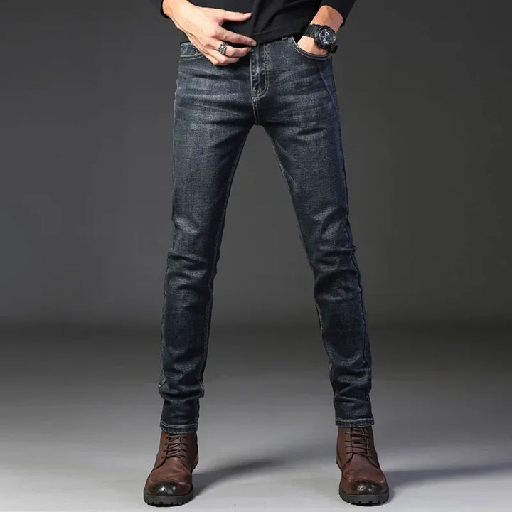 A person wearing dark slim-fit jeans crafted from stretch denim, a black long-sleeved top, brown lace-up boots, and a watch poses with hands in their pockets against a plain dark background.