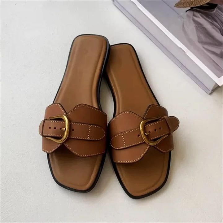 A pair of brown leather slides with wide straps, gold buckles, and white stitching rests on a light surface near the corner of a closed magazine.
