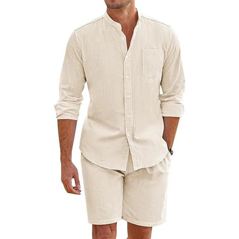 A man wearing a light beige, vintage stand collar shirt with a front pocket and matching baggy shorts. His sleeves are rolled up, and the shirt is partially unbuttoned for a casual, comfortable cotton linen outfit.