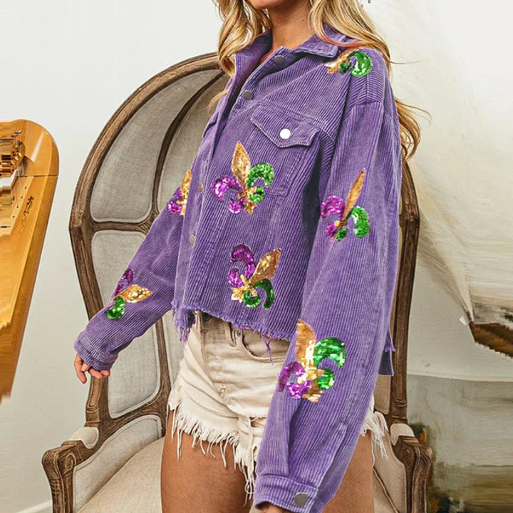 A woman wearing a cropped relaxed fit corduroy jacket with colorful sequin floral patches and distressed white denim shorts stands beside a vintage chair.