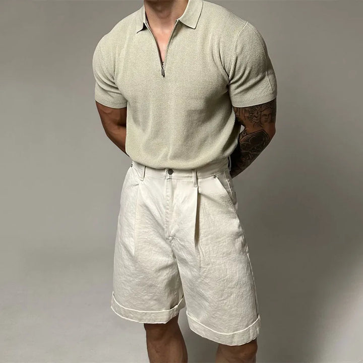 A man with muscular arms wears a light beige viscose polo with ribbed detailing and high-waisted, rolled-hem off-white shorts, standing with his hands behind his back against a plain background.