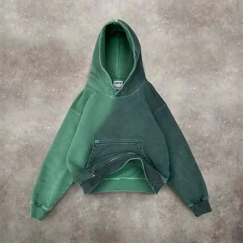 A green hooded sweatshirt with a front pocket and visible seams is laid flat on a textured, light brown surface. The hoodie, crafted from breathable fabric, appears slightly worn and has a relaxed, oversized fit.
