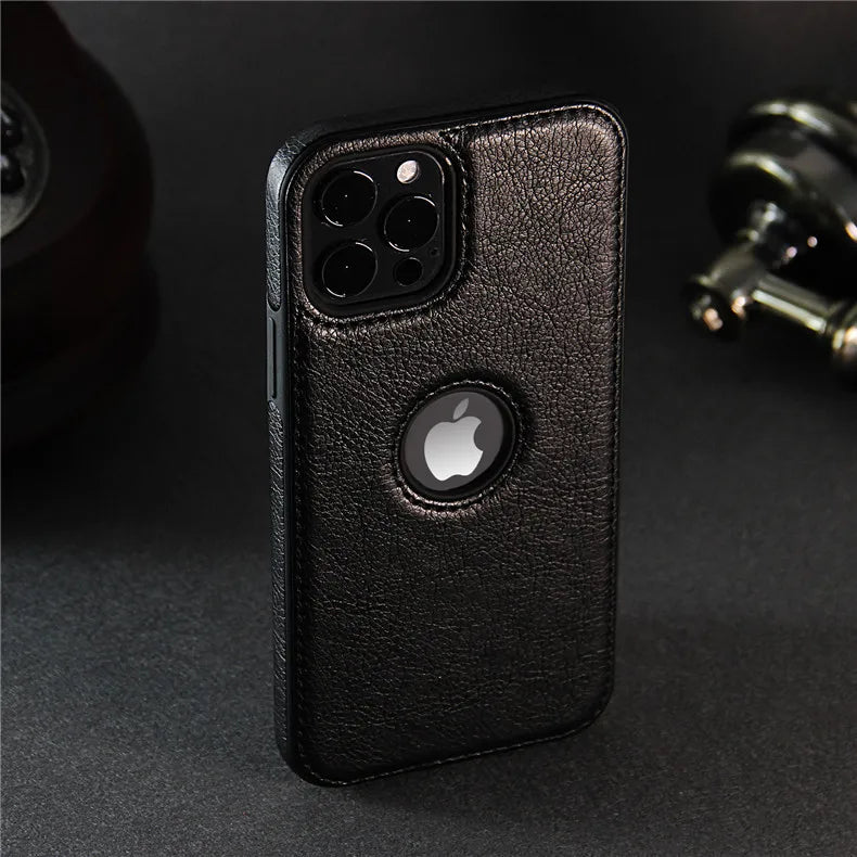 A luxury leather phone case in black, featuring a circular cutout for the Apple logo. This slim protective case stands upright on a dark surface, with blurred objects in the background and is wireless charging compatible.