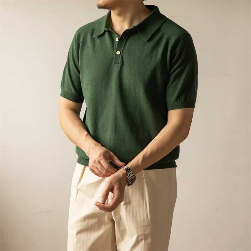 A person wearing a green classic style polo and light beige pants stands against a plain background, adjusting a watch on their wrist. Their face is not visible.