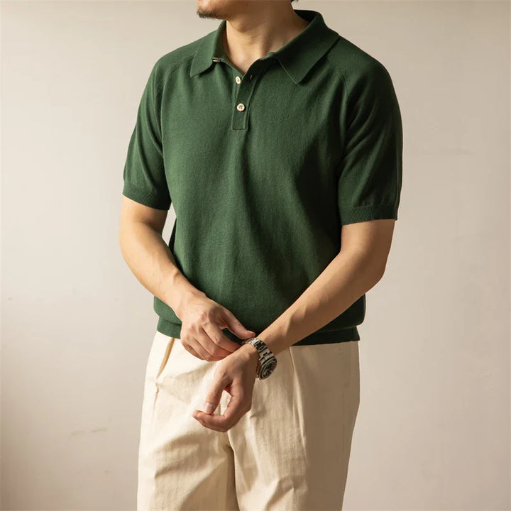 A person wearing a green classic style polo and light beige pants stands against a plain background, adjusting a watch on their wrist. Their face is not visible.
