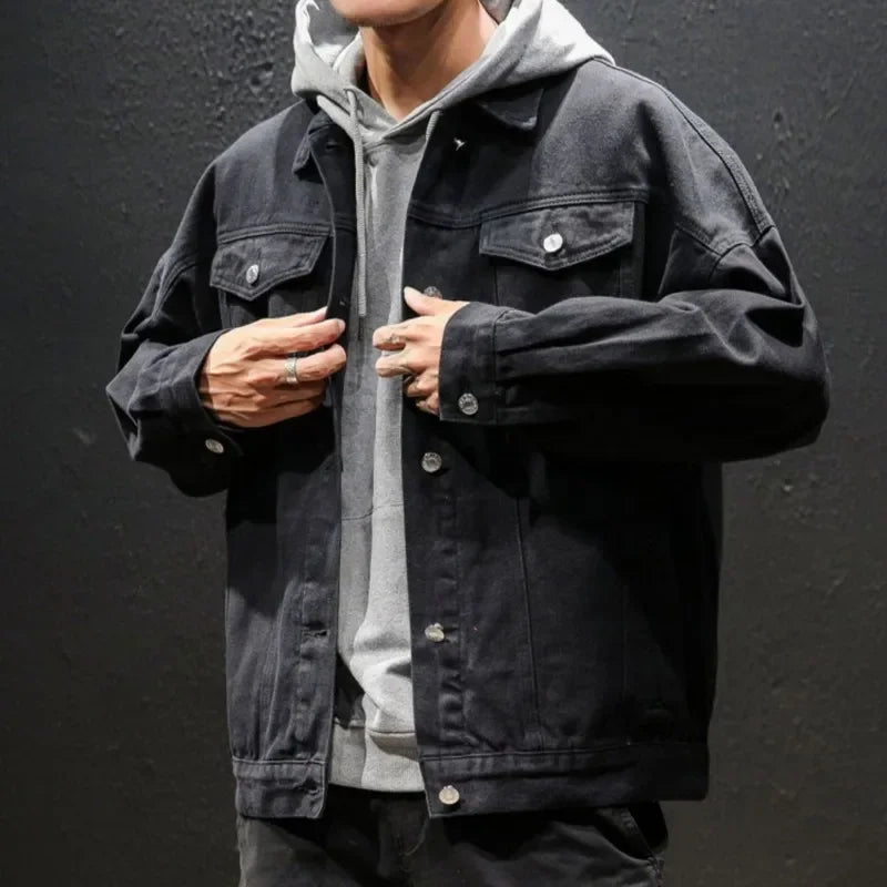 A person in a gray hoodie layered under a black premium denim jacket stands against a dark textured wall, adjusting the button-down jacket with both hands near the buttons. Their face is partially out of frame.
