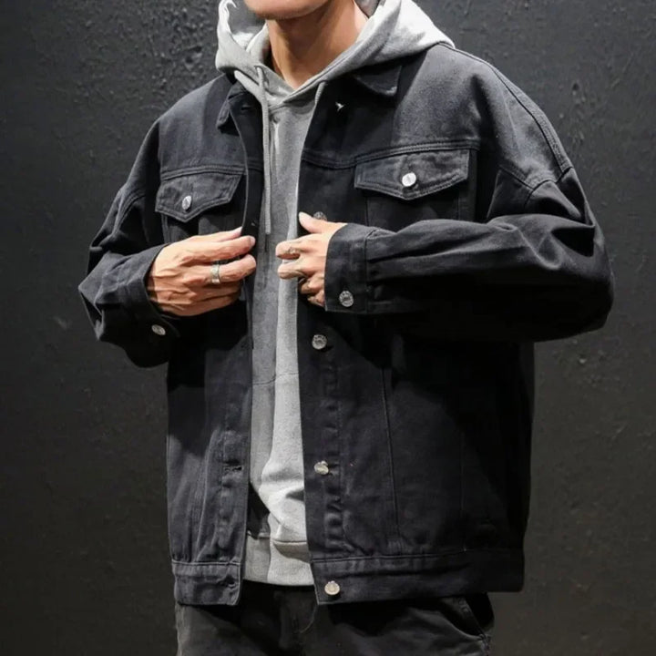 A person in a gray hoodie layered under a black premium denim jacket stands against a dark textured wall, adjusting the button-down jacket with both hands near the buttons. Their face is partially out of frame.