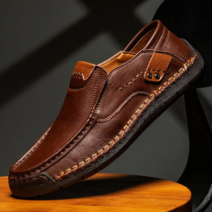 A brown leather loafer with detailed stitching and tan accents showcases traditional craftsmanship, resting on a wooden surface against a dark background.