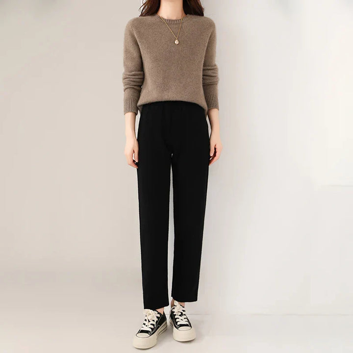 A person stands against a plain background wearing a brown sweater with wool cashmere lining, black pants with a slim tailored leg, black-and-white sneakers, and layered necklaces. Their hands are relaxed at their sides and their face is not visible.