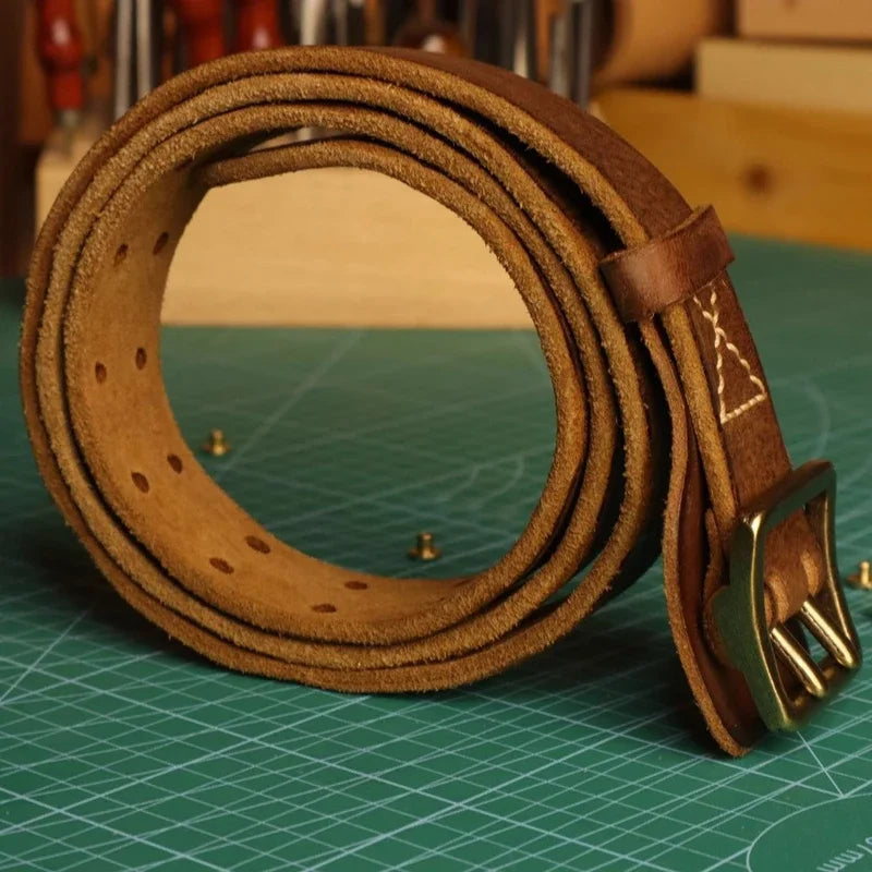 A rolled-up cowhide belt with a brass buckle sits on a green cutting mat, exuding a rugged aesthetic, with tools and a wooden background visible behind it.