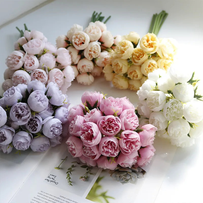 Several bouquets of peony flowers in pastel colors—pink, white, yellow, lavender, and cream—are arranged on a white surface with realistic faux petals, creating a soft, elegant display. Some text and a plant are partially visible beneath them.