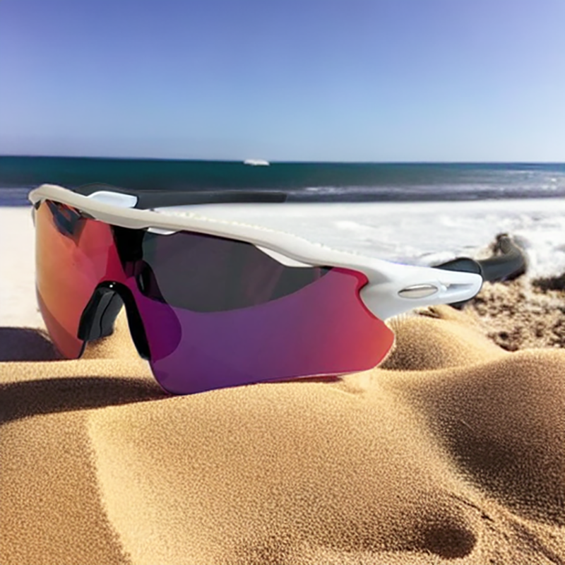 A pair of white-framed, mirrored Polarized Cycling Sunglasses with UV Protection resting on sandy beach with the ocean and blue sky in the background.