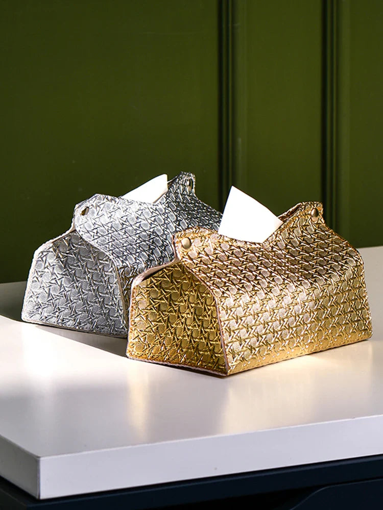 Two decorative tissue holders, including a Glymmer Tissue Box in metallic gold and a silver geometric tissue box, sit side by side on a white table against a green background. A tissue is partially pulled out from the gold holder.