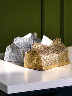 Two decorative tissue holders, including a Glymmer Tissue Box in metallic gold and a silver geometric tissue box, sit side by side on a white table against a green background. A tissue is partially pulled out from the gold holder.