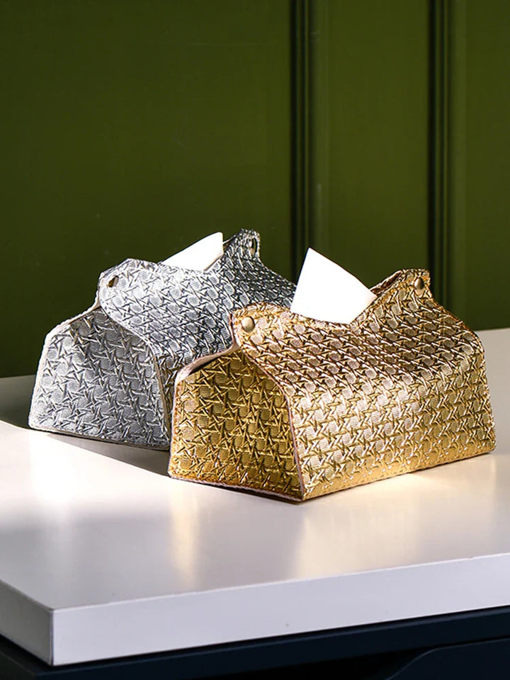 Two decorative tissue holders, including a Glymmer Tissue Box in metallic gold and a silver geometric tissue box, sit side by side on a white table against a green background. A tissue is partially pulled out from the gold holder.