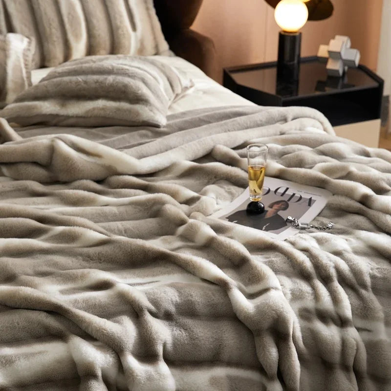 A cozy bed with a soft, striped, gray and white faux rabbit fur blanket, matching pillows, a magazine, eyeglasses, and a glass of drink on top. A modern nightstand with a lamp and decorative objects sits beside the bed.