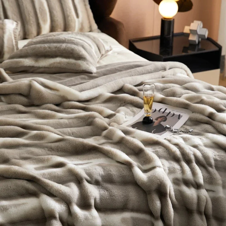 A cozy bed with a soft, striped, gray and white faux rabbit fur blanket, matching pillows, a magazine, eyeglasses, and a glass of drink on top. A modern nightstand with a lamp and decorative objects sits beside the bed.