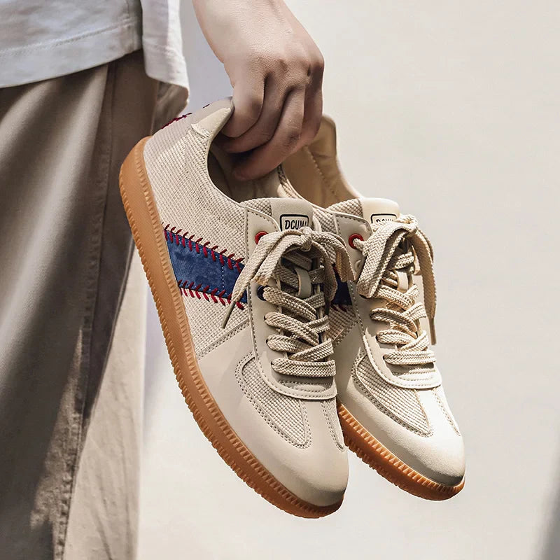 A person holding a pair of beige baseball inspired shoes with gum soles, blue and red side stripes, and beige laces. Only the hand and lower part of the persons clothing are visible.