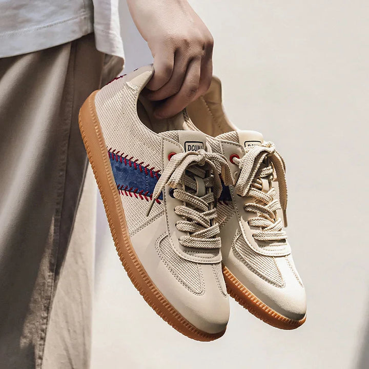 A person holding a pair of beige baseball inspired shoes with gum soles, blue and red side stripes, and beige laces. Only the hand and lower part of the persons clothing are visible.