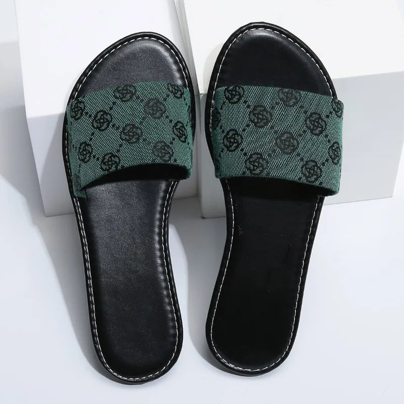 A pair of black open-toe slide sandals with green fabric straps featuring a repeating black pattern, these modern slippers add a touch of British style and are perfect for summer days, displayed on a white surface.