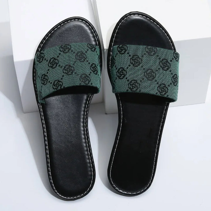 A pair of black open-toe slide sandals with green fabric straps featuring a repeating black pattern, these modern slippers add a touch of British style and are perfect for summer days, displayed on a white surface.