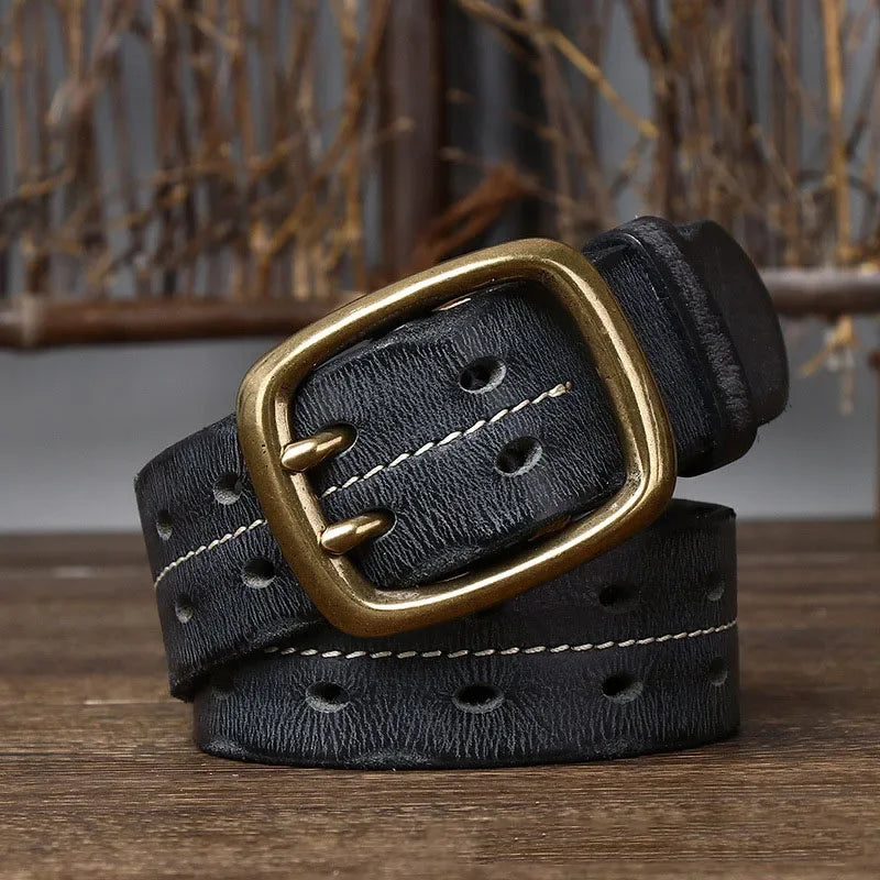 A genuine leather belt with double rows of white stitching and a large, rectangular brass buckle is coiled on a wooden surface, highlighting heritage craftsmanship against a rustic backdrop of brown branches.