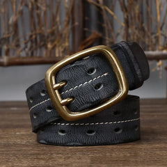 A genuine leather belt with double rows of white stitching and a large, rectangular brass buckle is coiled on a wooden surface, highlighting heritage craftsmanship against a rustic backdrop of brown branches.