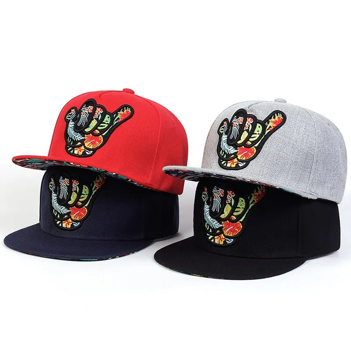 Four snapback hats with a colorful embroidered shaka hand symbol on the front. Each Hawaiian-inspired cap comes in red, gray, navy blue, or black, featuring a vibrant floral pattern under the brim for that tropical snapback vibe.