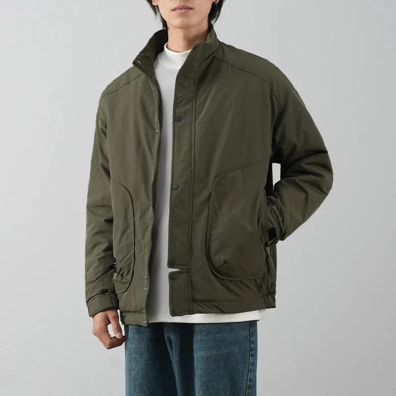 A person wearing a white t-shirt, blue jeans, and an olive green minimalist jacket with snap buttons and luxurious cotton lining stands against a plain light gray background.