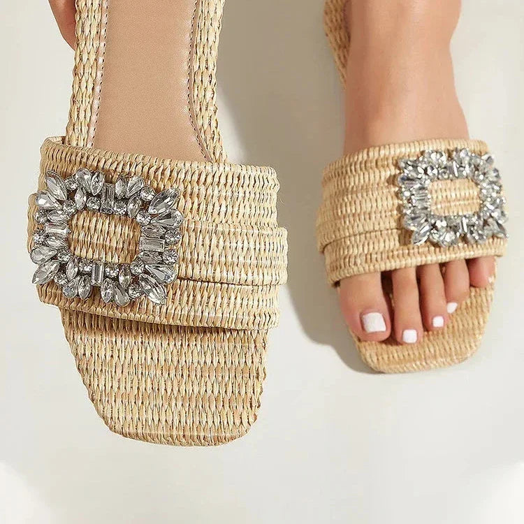 A close-up of feet in handwoven cane slip-on flats with square toes, adorned with rectangular crystal buckles. The person has neatly manicured toenails painted white.