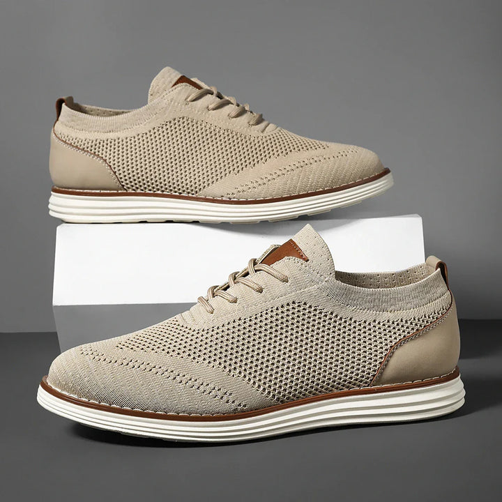 A pair of beige breathable knit sneakers with white soles and brown accents, displayed against a gray background, with one shoe resting on a white box and the other positioned in front, perfect for casual style.