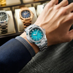 A person wearing a stainless steel wristwatch with a turquoise blue dial, dressed in a navy suit, displays the watch over a dark textured surface with several other luxury watches in a display case in the background.
