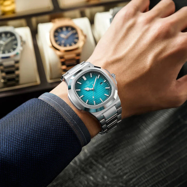 A person wearing a stainless steel wristwatch with a turquoise blue dial, dressed in a navy suit, displays the watch over a dark textured surface with several other luxury watches in a display case in the background.