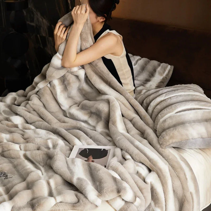 A woman sits on a bed covered with a soft, plush blanket in gray-and-white stripes, partially hiding her face. A pillow and an open magazine rest nearby, adding to the cozy scene.