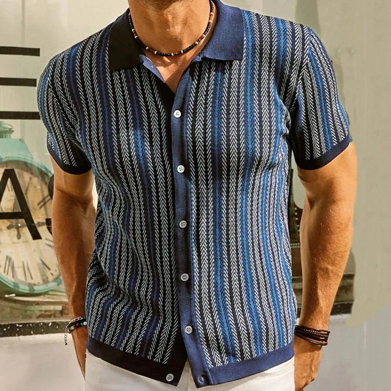 A man wearing a short-sleeved, button-up shirt with a blue and black jacquard herringbone pattern, white buttons, and a black collar, stands in front of a window with part of a clock visible in the background.