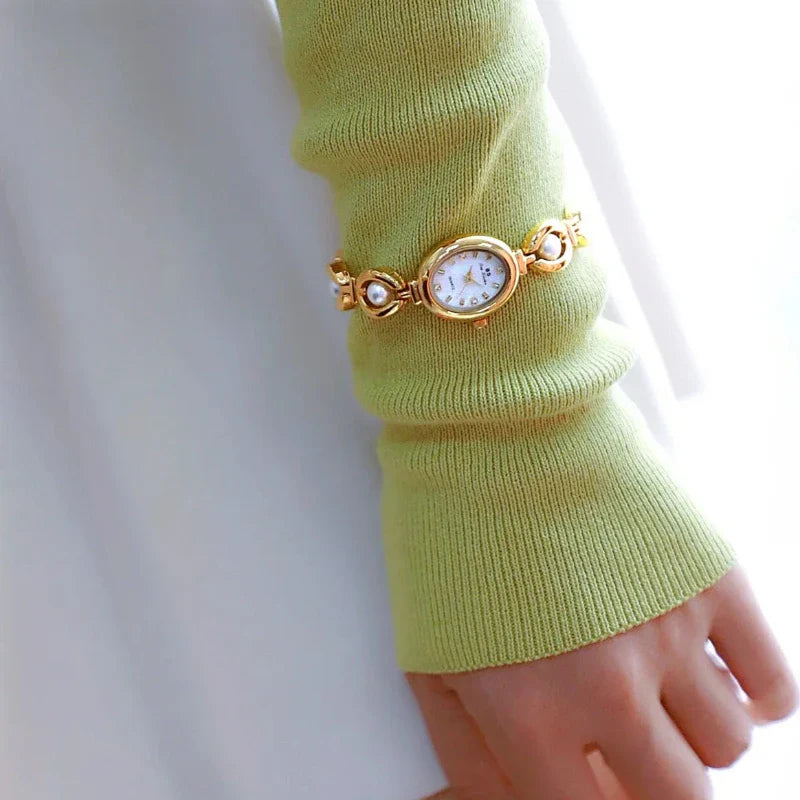 A person wearing a light green, long-sleeved sweater has a gold wristwatch with pearl accents over the sleeve near the wrist. The softly lit, mostly white background enhances the elegance of the pearl accents on the watch.