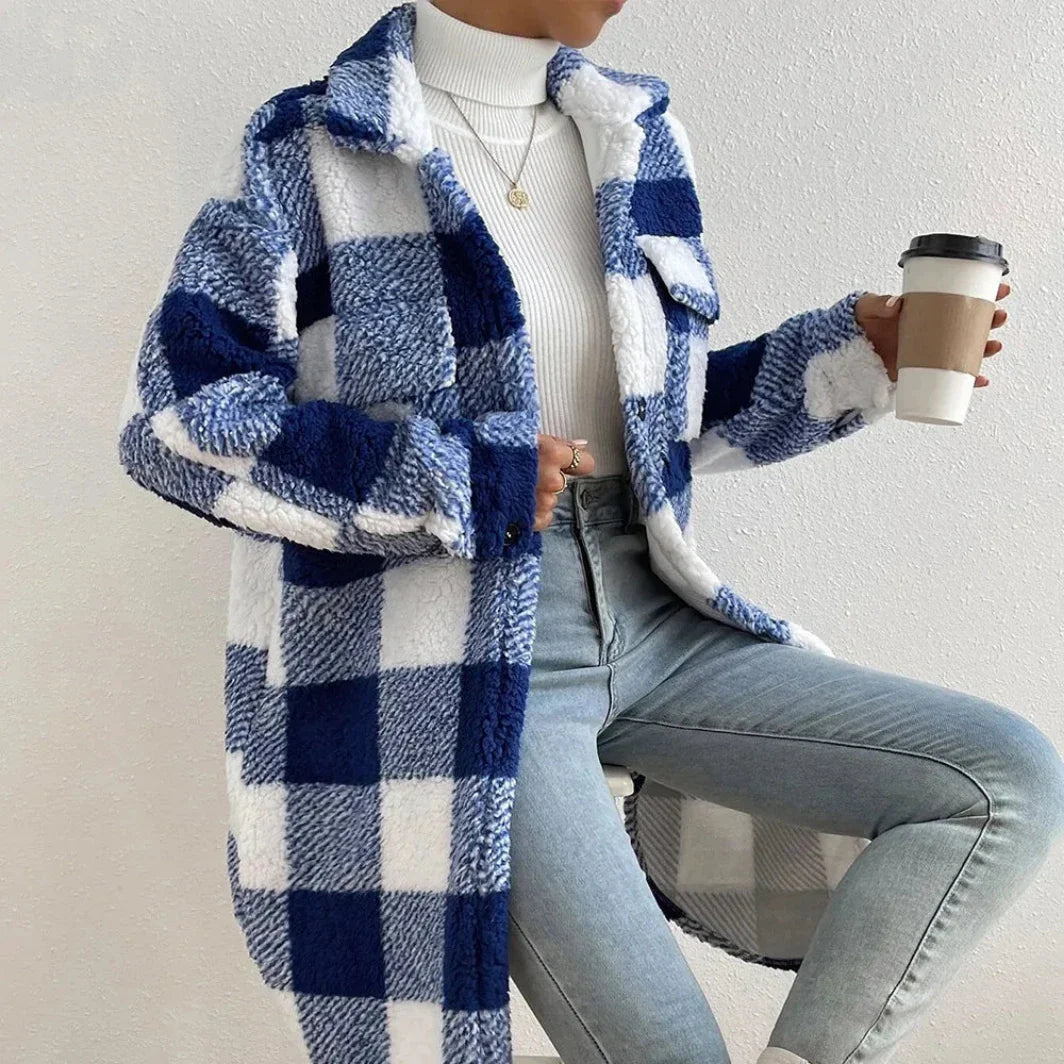 A person wearing a blue and white plaid Sherpa coat with an open lapel, a white turtleneck, and light blue jeans holds a coffee cup and sits on a stool against a light-colored wall.