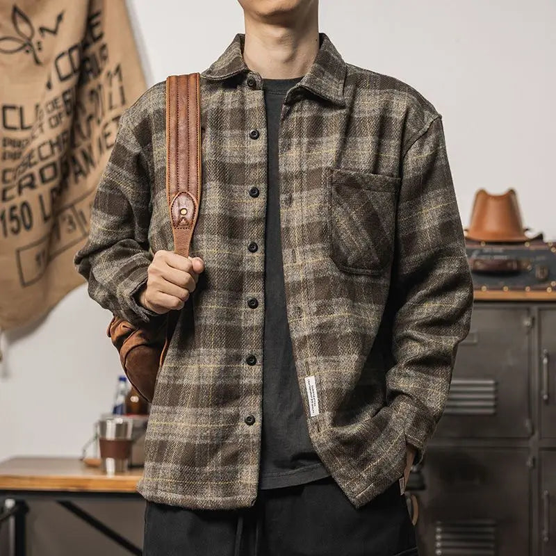 A person wearing a brown and gray plaid relaxed fit button-up shirt with a front pocket, holding a brown leather strap over their shoulder, standing indoors near a metal cabinet and a hanging canvas bag.