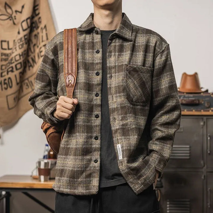 A person wearing a brown and gray plaid relaxed fit button-up shirt with a front pocket, holding a brown leather strap over their shoulder, standing indoors near a metal cabinet and a hanging canvas bag.