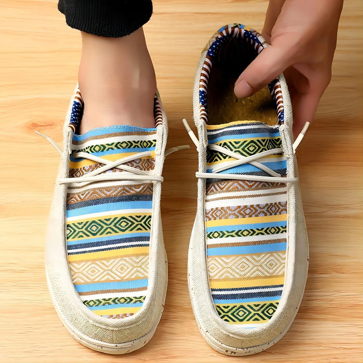 A person puts on slip-on shoes with a woven upper, white soles, and colorful geometric patterns in blue, yellow, and green on a wooden floor.