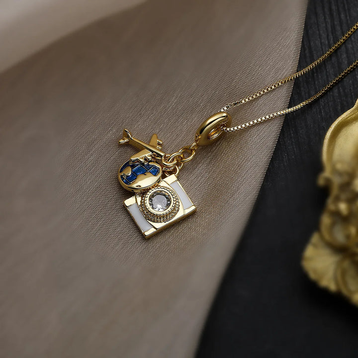 A gold adventure necklace with a delicate chain, featuring a globe charm in blue and gold plus a camera charm with a clear gem lens, resting on a beige and black fabric background.
