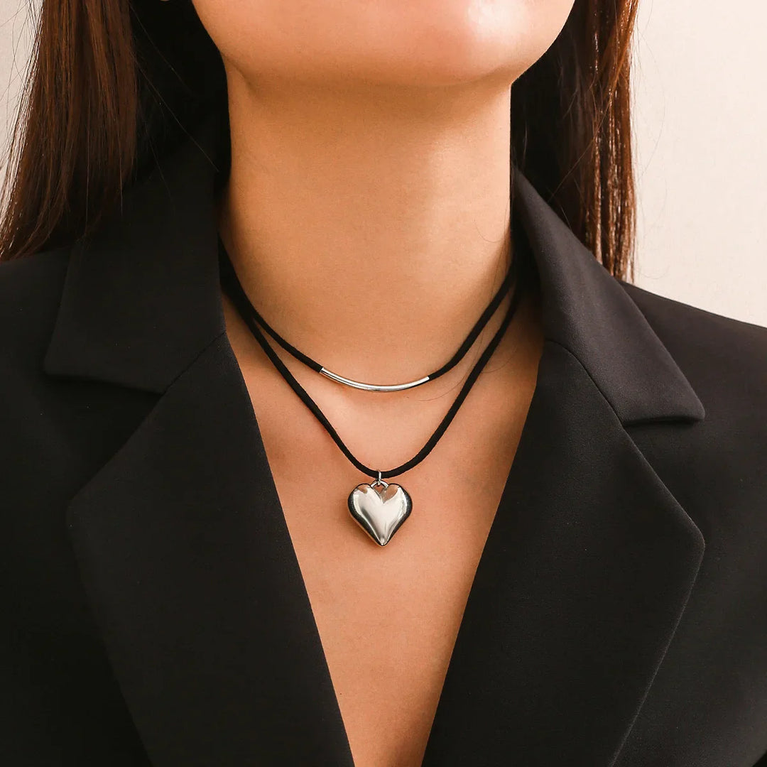 A woman wears a black blazer and two layered necklaces: a classic velvet choker and an adjustable cord featuring a vintage heart pendant. Only her neck and lower face are visible.
