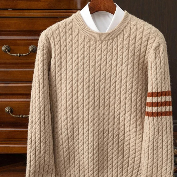 A beige cable-knit cashmere sweater with brown stripes on the left sleeve, featuring ribbed cuffs, displayed on a wooden hanger in front of a wooden dresser.