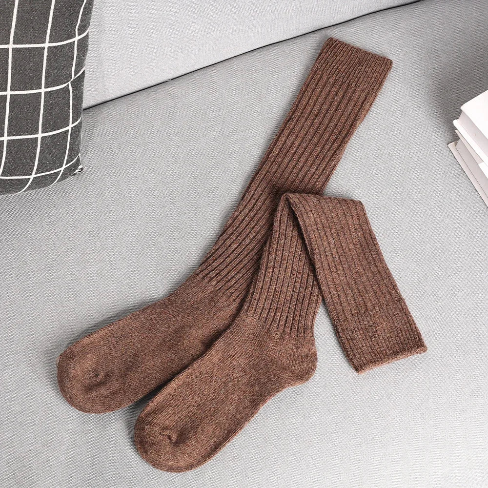 A pair of long, ribbed brown socks made from a cozy Cashmere cotton blend is laid out neatly on a light gray couch next to a white-and-black checkered pillow and a stack of white books.