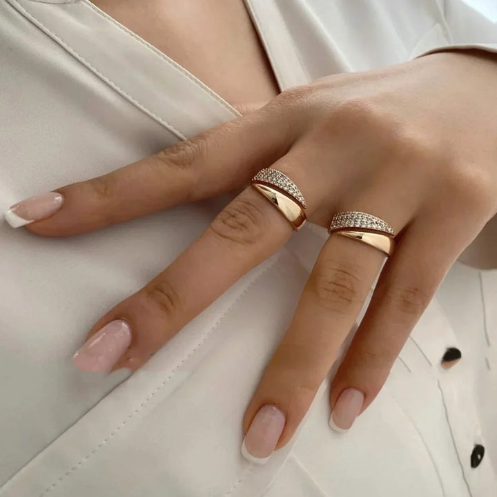 A hand with light pink manicured nails wears two gold rings—a twisted design and one with pavé diamond accents—on the middle and ring fingers, resting against a white fabric background.